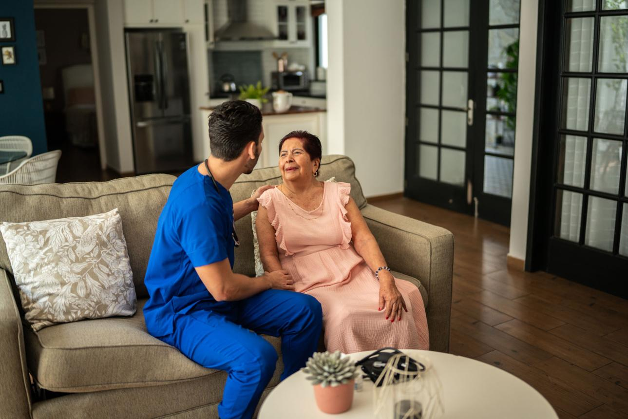 Soignant en blouse bleue échangeant avec une dame âgée souriante assise sur un canapé — illustration démence et incontinence